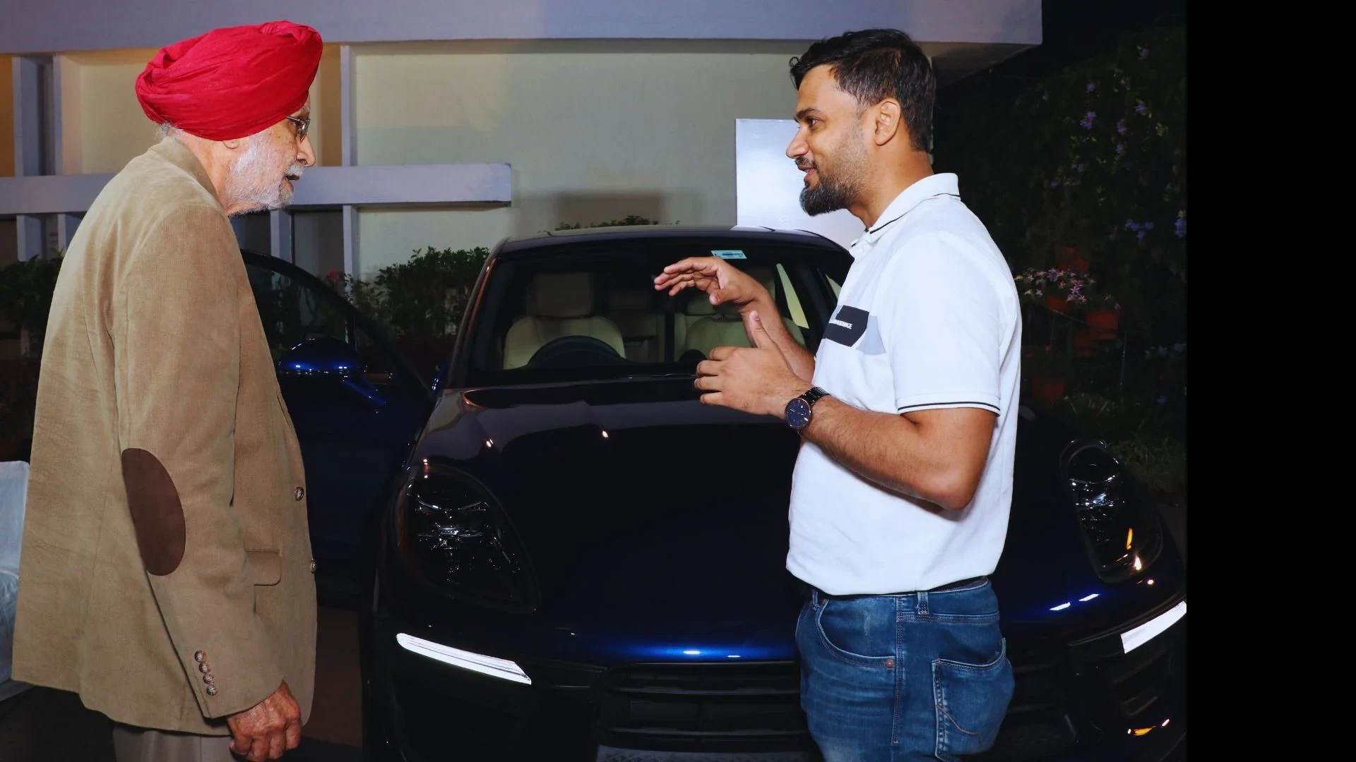 A man in a red turban and a man in a white polo shirt talking beside the blue Porsche at the event