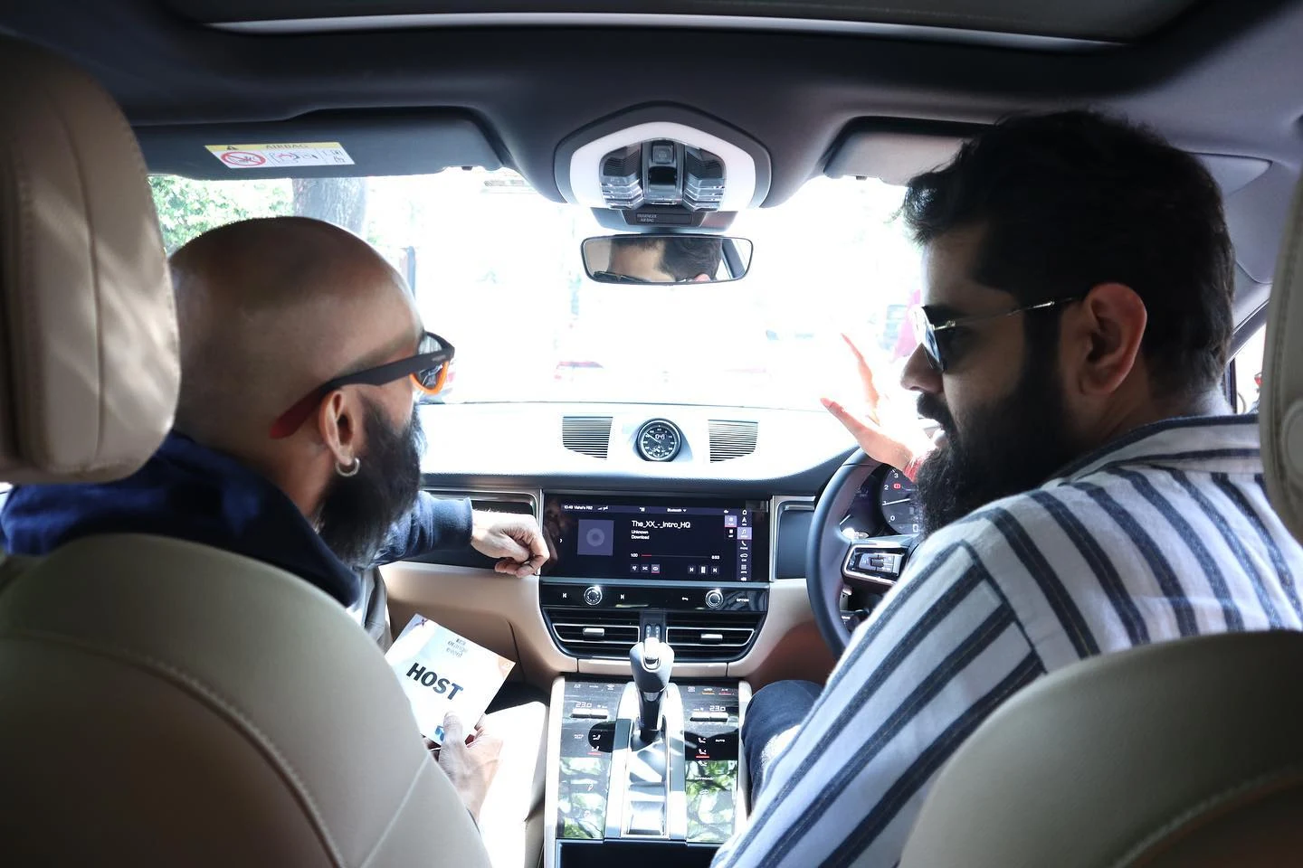 Aditya in the driver’s seat turning toward the creator in the front passenger seat of the Porsche, both visible from the rear seat