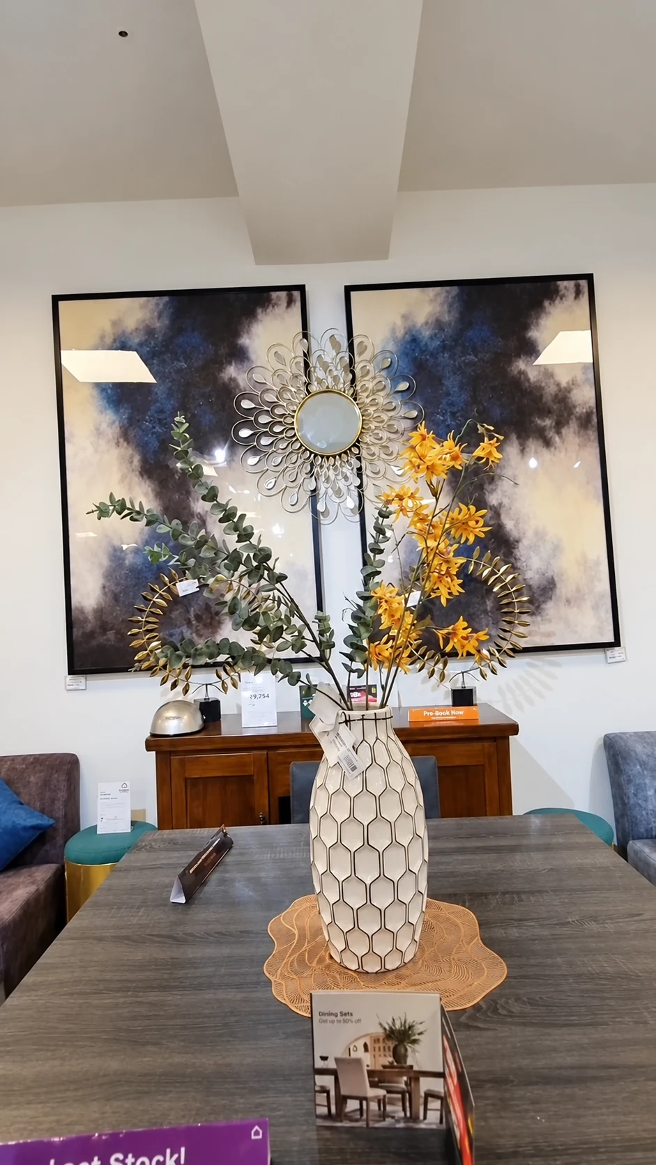 A dining table with a patterned vase, decorative branches and product information, with artwork behind it