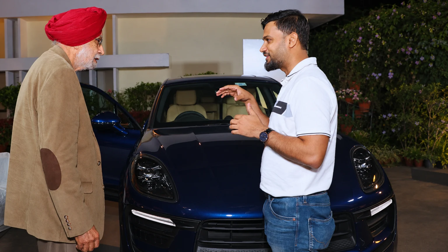 Two men in conversation beside a dark blue Porsche, one explaining and the other listening