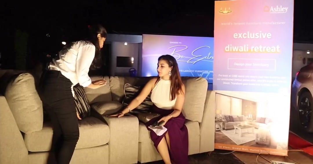 A guest seated in an Ashley recliner, talking with another woman beside an Ashley event display