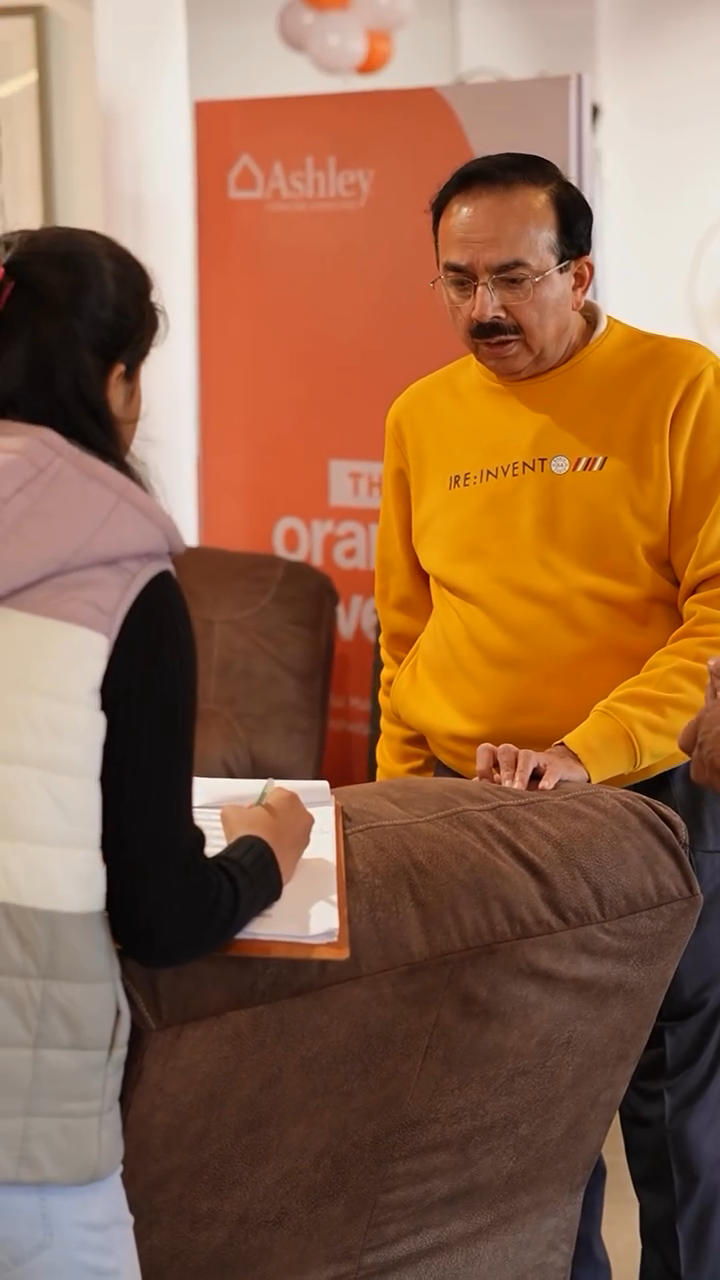 A visitor in a yellow sweater speaking with a woman holding a clipboard beside a sofa at the Orange Event