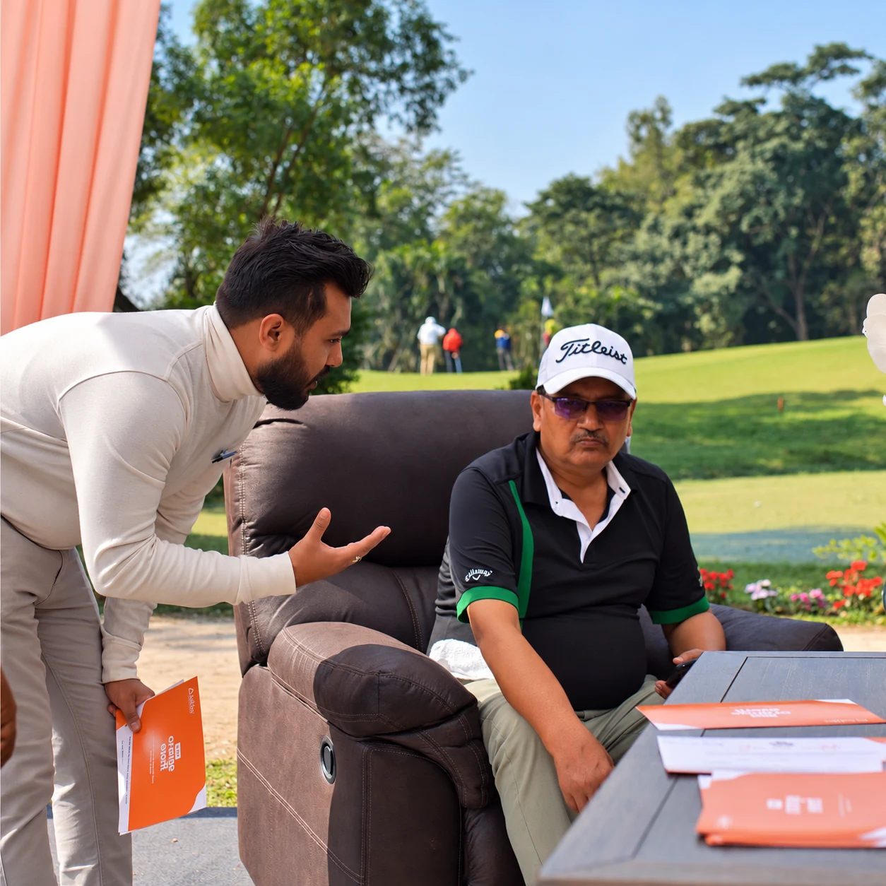 An Ashley team member speaking with a golfer seated in a recliner, with Orange Event invitations beside them