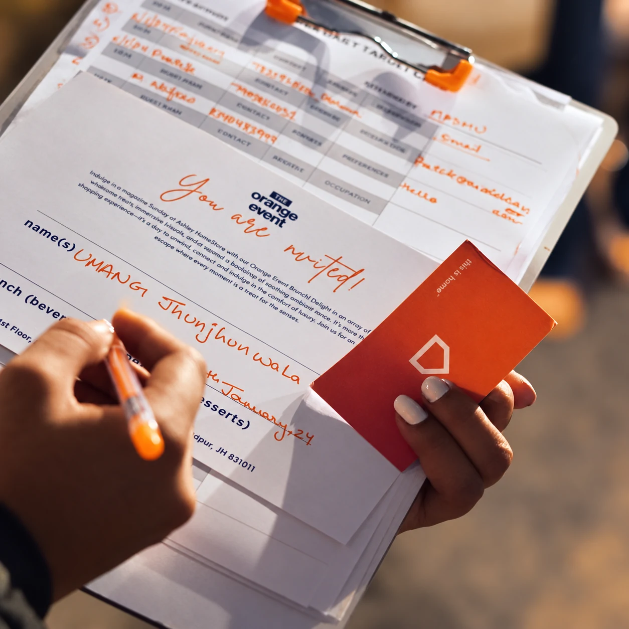 An Orange Event invitation being filled in by hand beside an orange Ashley card